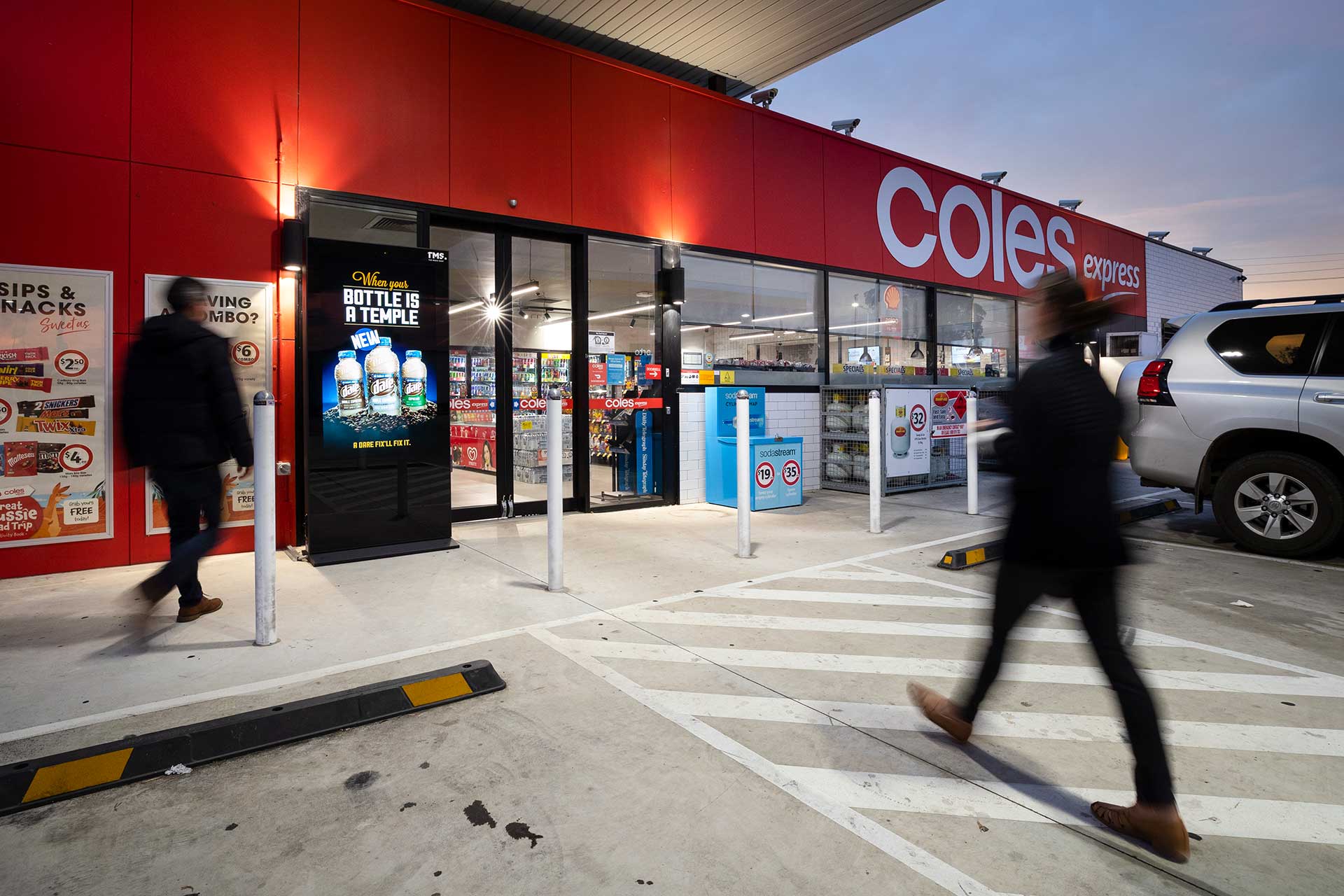 Digital advertising banner outside Coles Express service station entrance with people entering